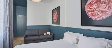 Una habitación de hotel con una cama, un sofá gris, una obra de arte floral grande y una lámpara de techo blanca.