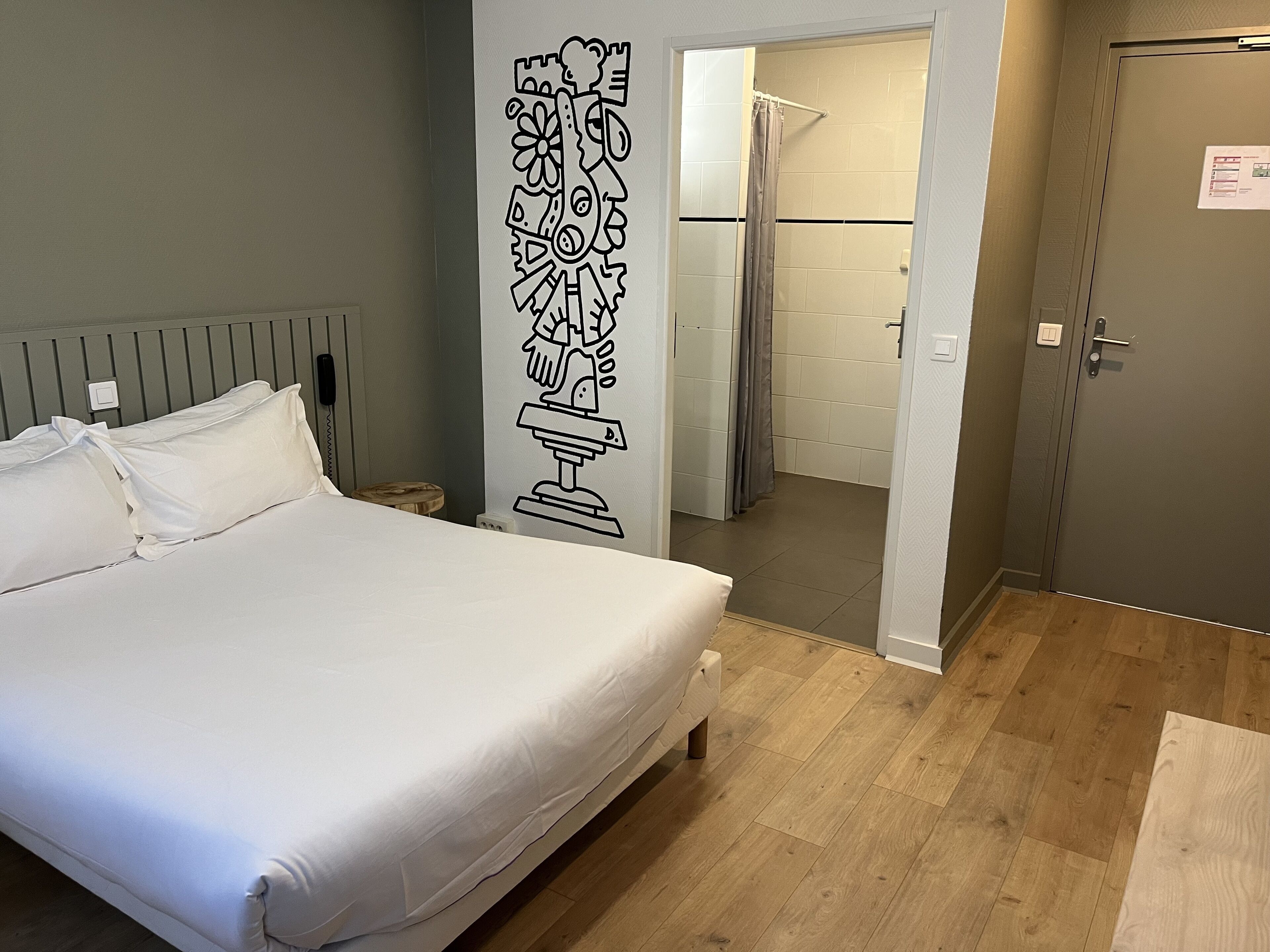 A bedroom with a bed, a wall-mounted artwork, a wooden floor, and a bathroom visible through an open door.