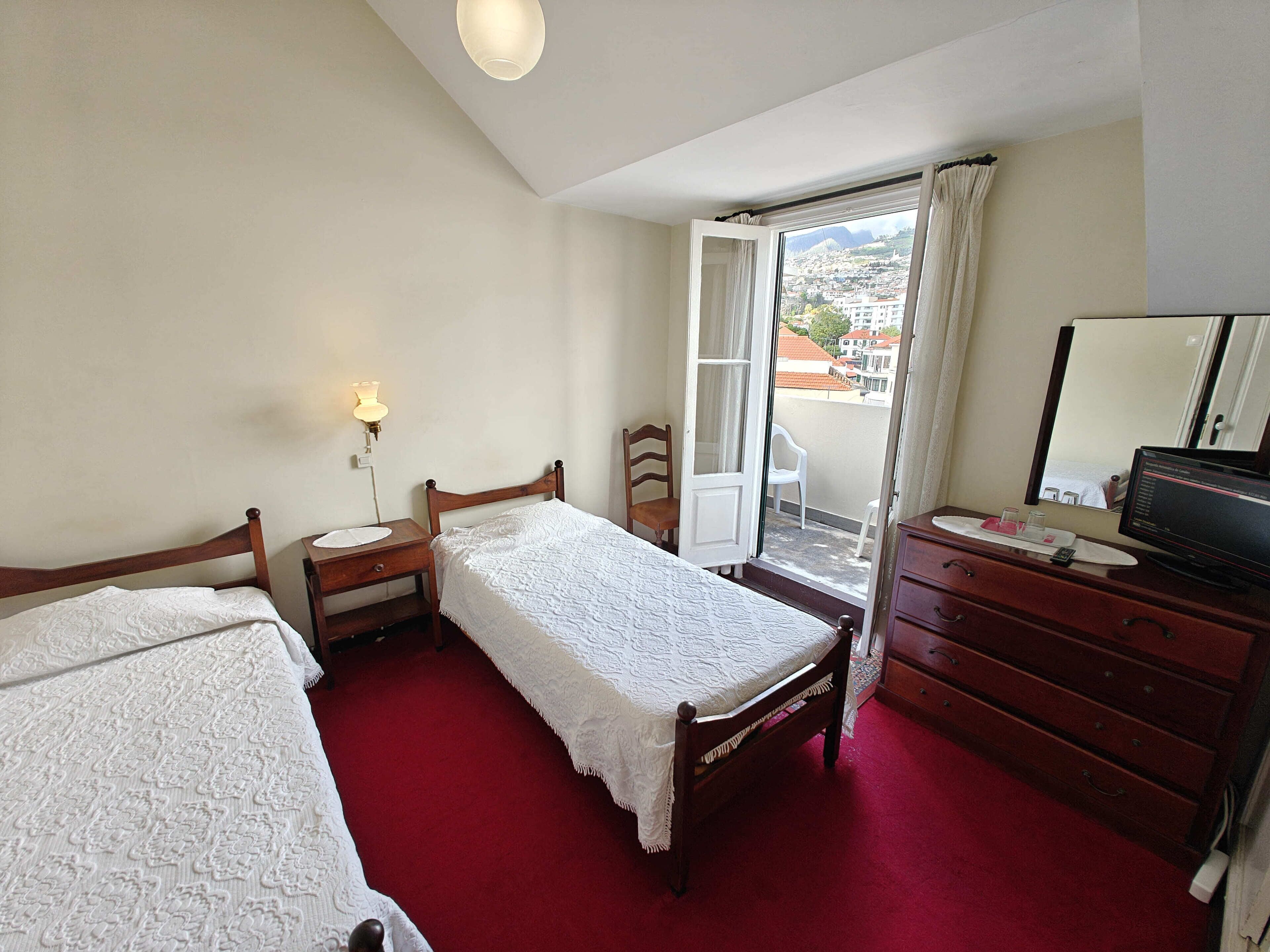 A bedroom with two single beds, a wooden dresser, a desk with a lamp, and a sliding glass door to a balcony.