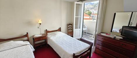 A bedroom with two single beds, a wooden dresser, a desk with a lamp, and a sliding glass door to a balcony.