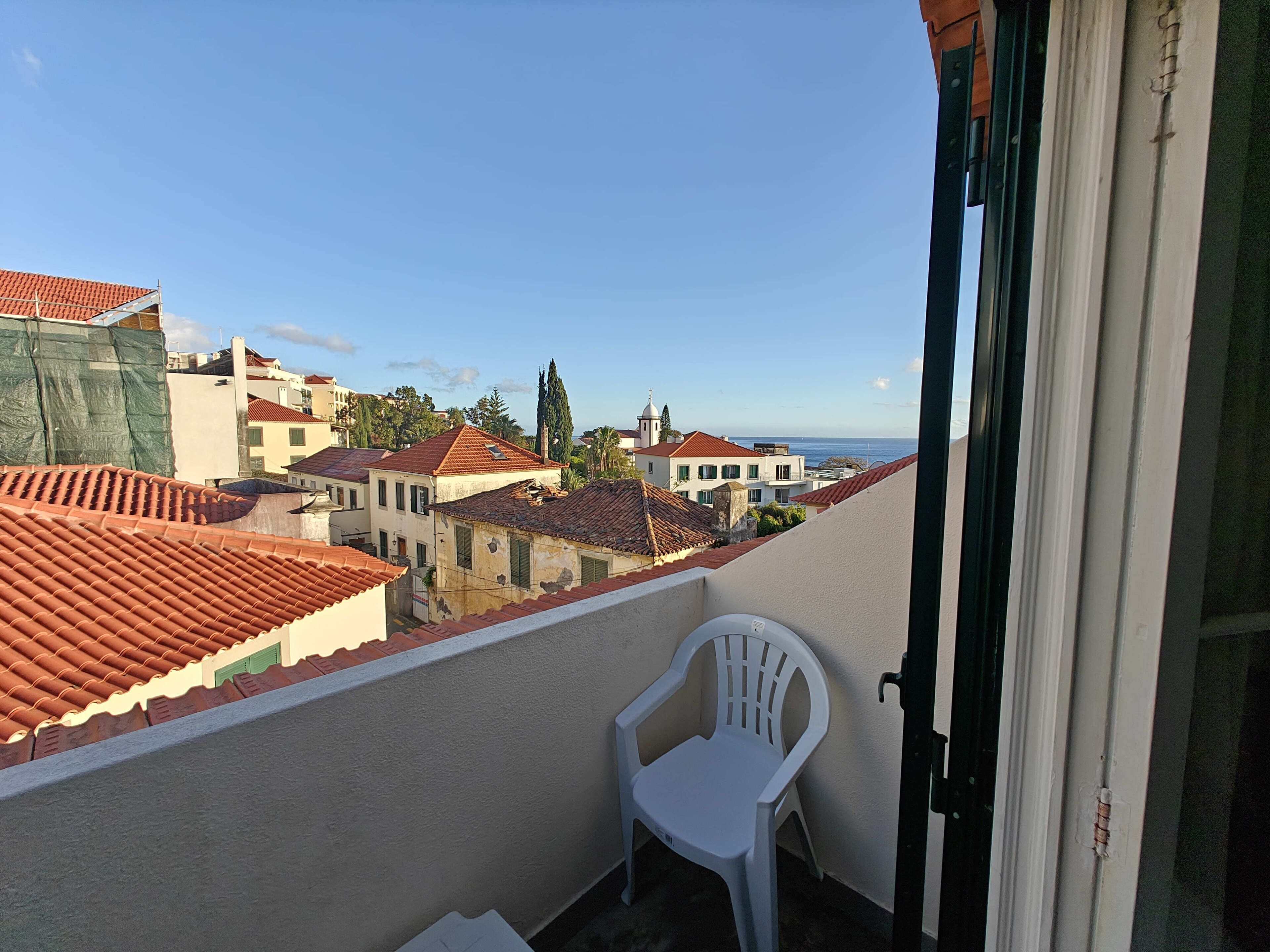 Uma varanda com uma cadeira de plástico branca, telhado de azulejos e vista para uma cidade costeira.