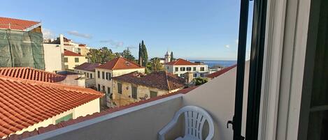Uma varanda com uma cadeira de plástico branca, telhado de azulejos e vista para uma cidade costeira.