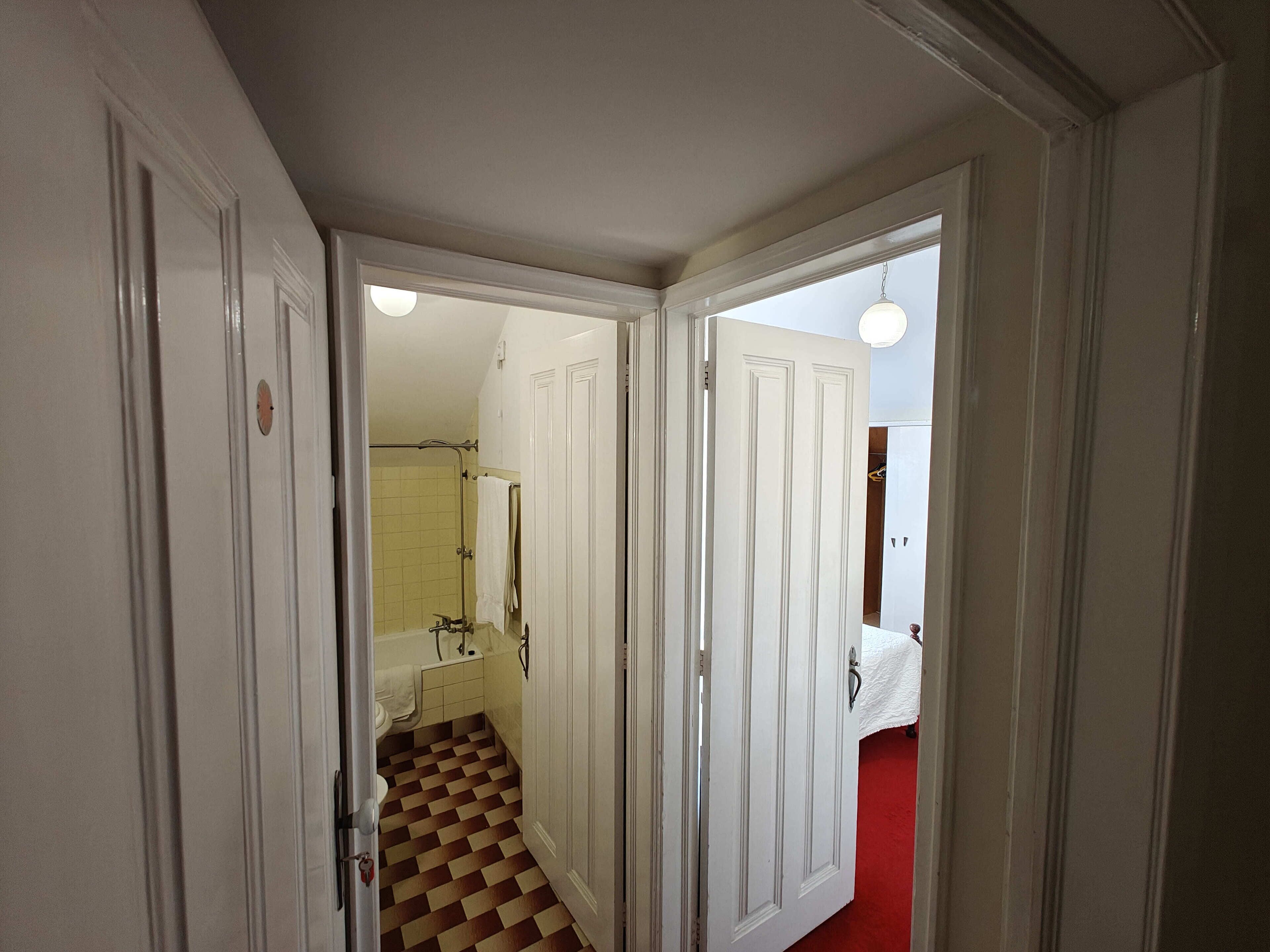 Un couloir mène à une salle de bain dont le sol est à carreaux, les murs sont blancs et l’espace de la douche est bien visible.
