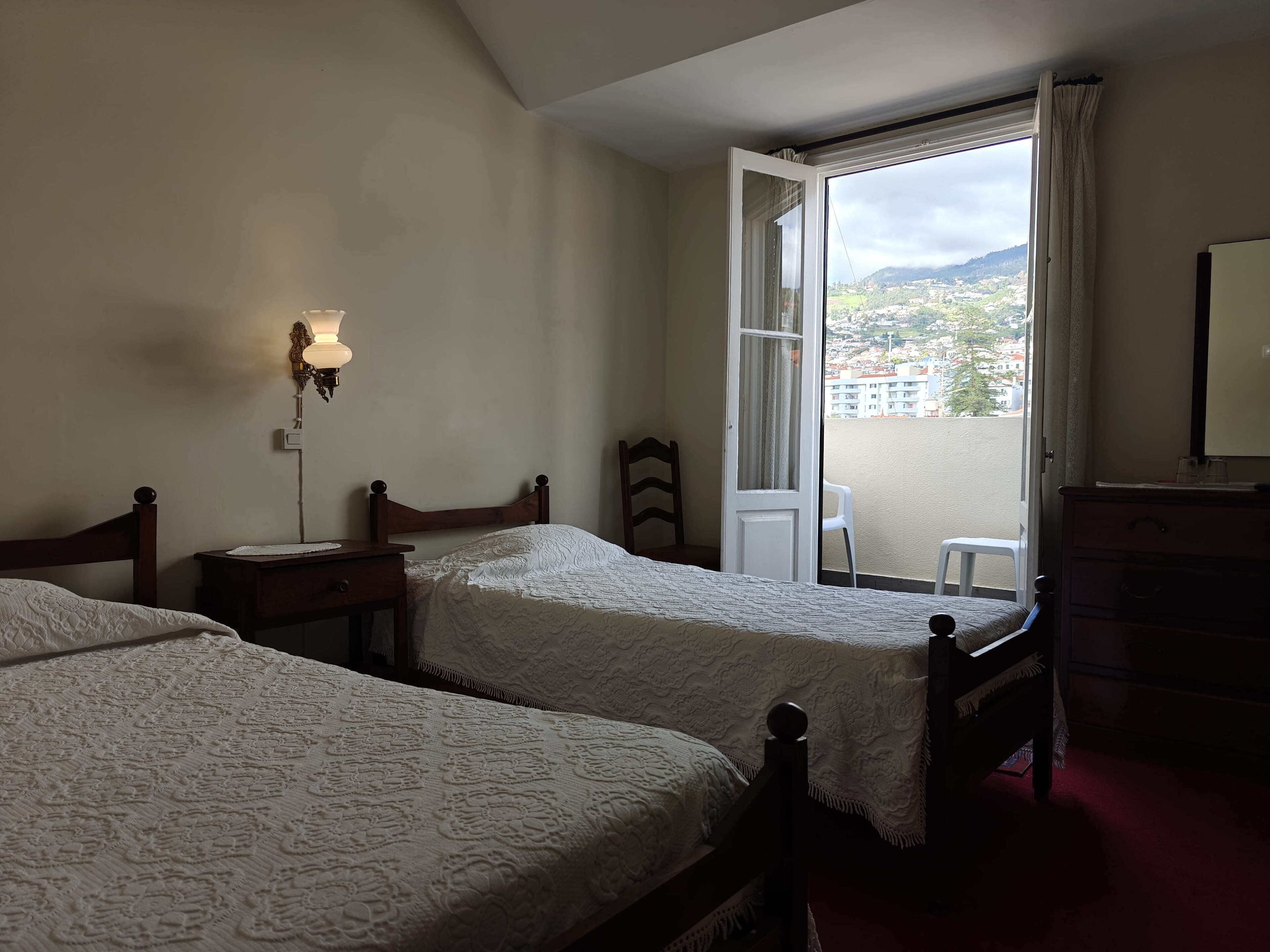 Une chambre d’hôtel avec deux lits, une table de chevet, une commode, une fenêtre donnant sur un paysage urbain et une lampe fixée au mur.