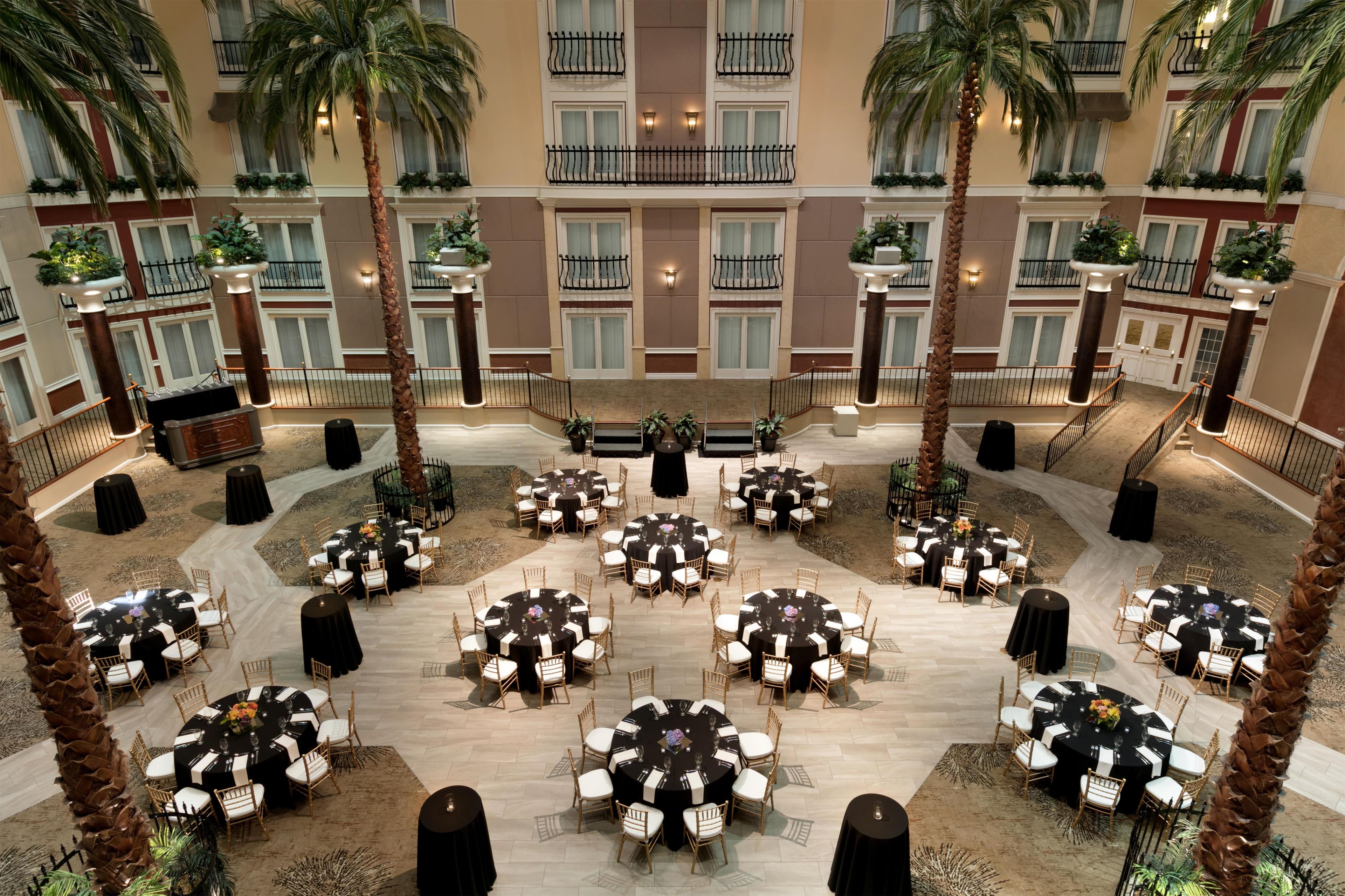 A hotel atrium with multiple round tables, palm trees, and a central staircase.
