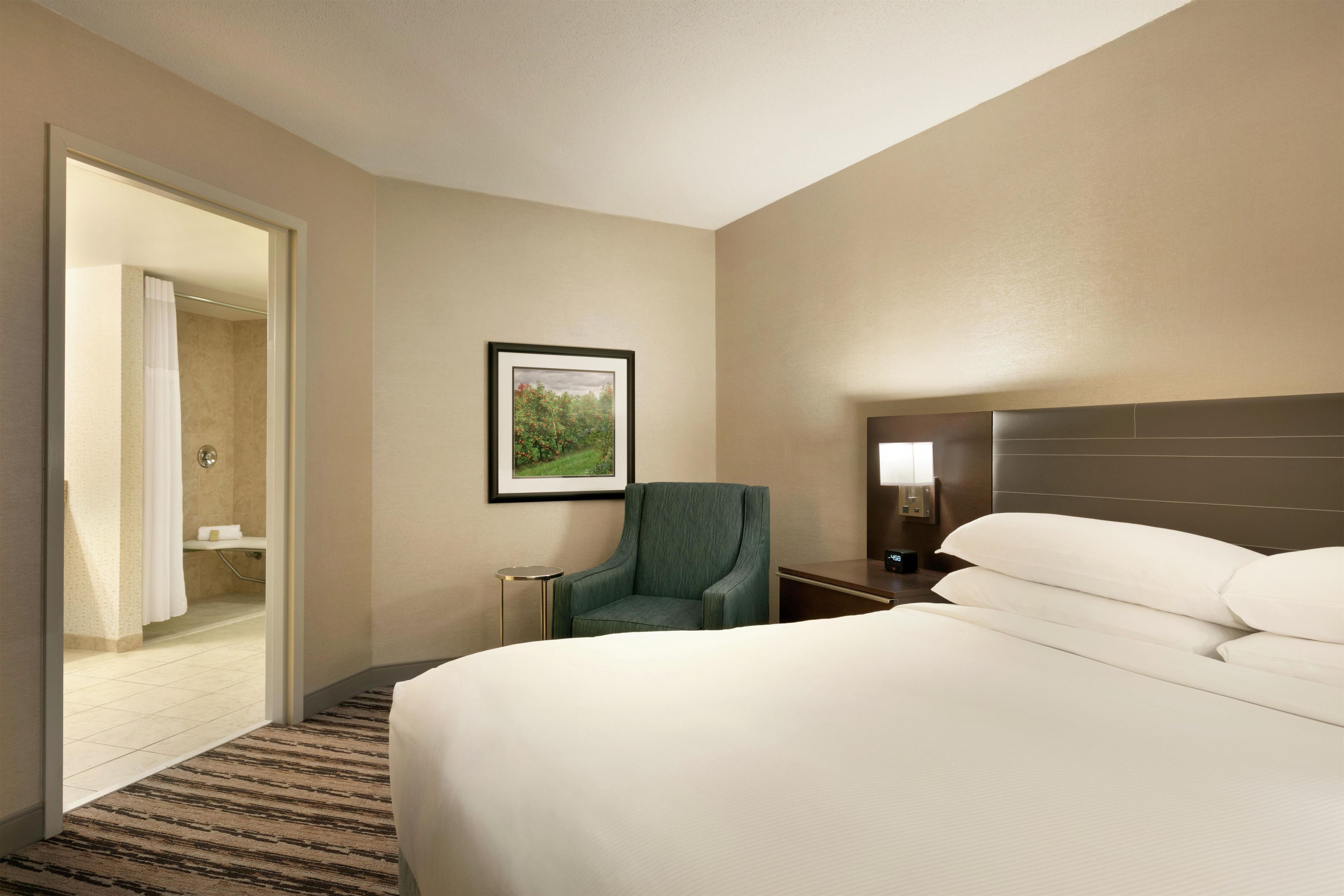 A hotel room with a bed, a chair, a bedside table, a nightstand, a lamp, and a framed picture on the wall.