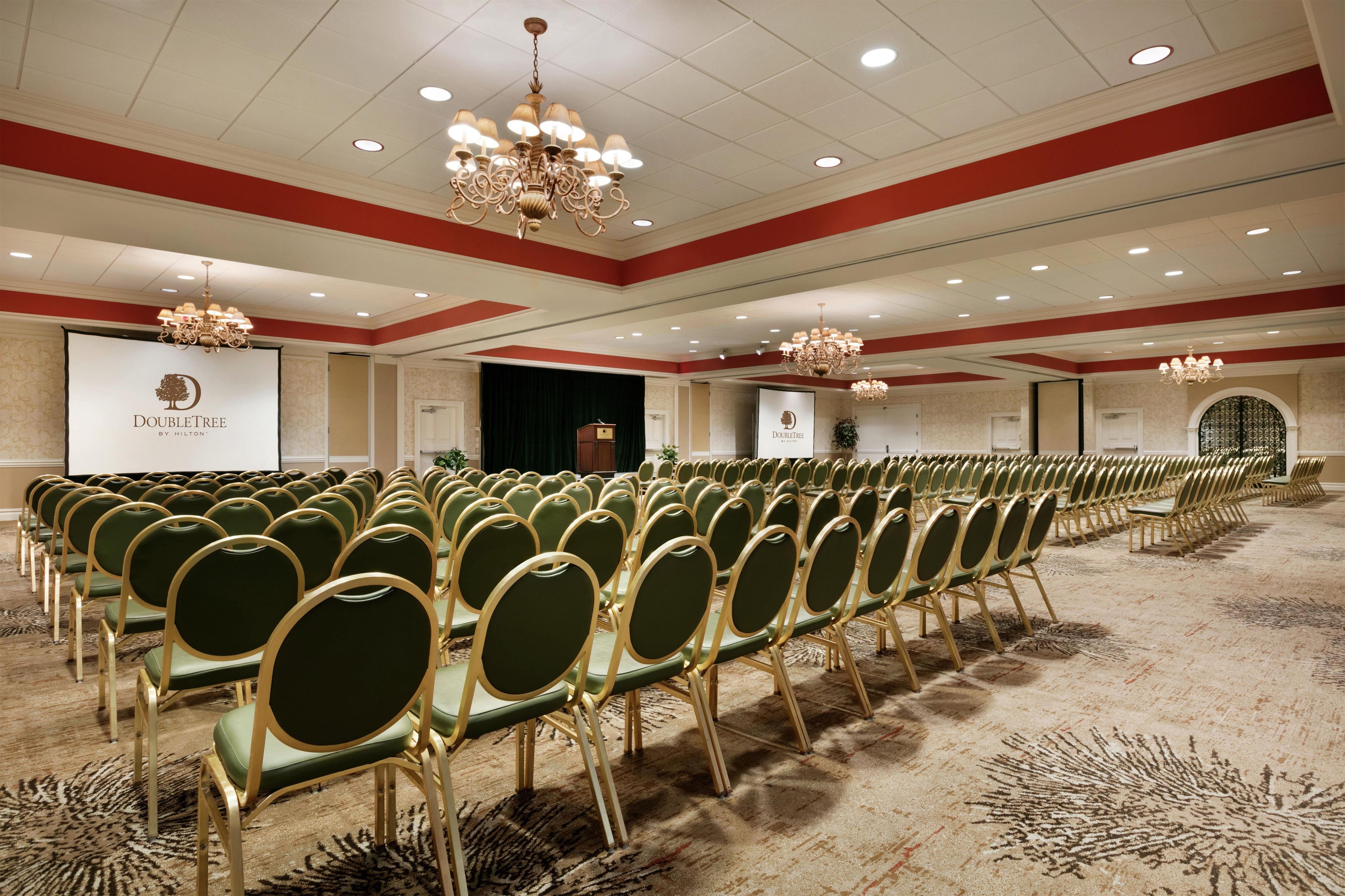 A conference room with rows of green chairs, a stage with a projector screen, and chandeliers.