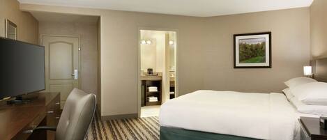 Habitación de hotel con cama, escritorio, silla, televisor y baño visible a través de una puerta abierta.