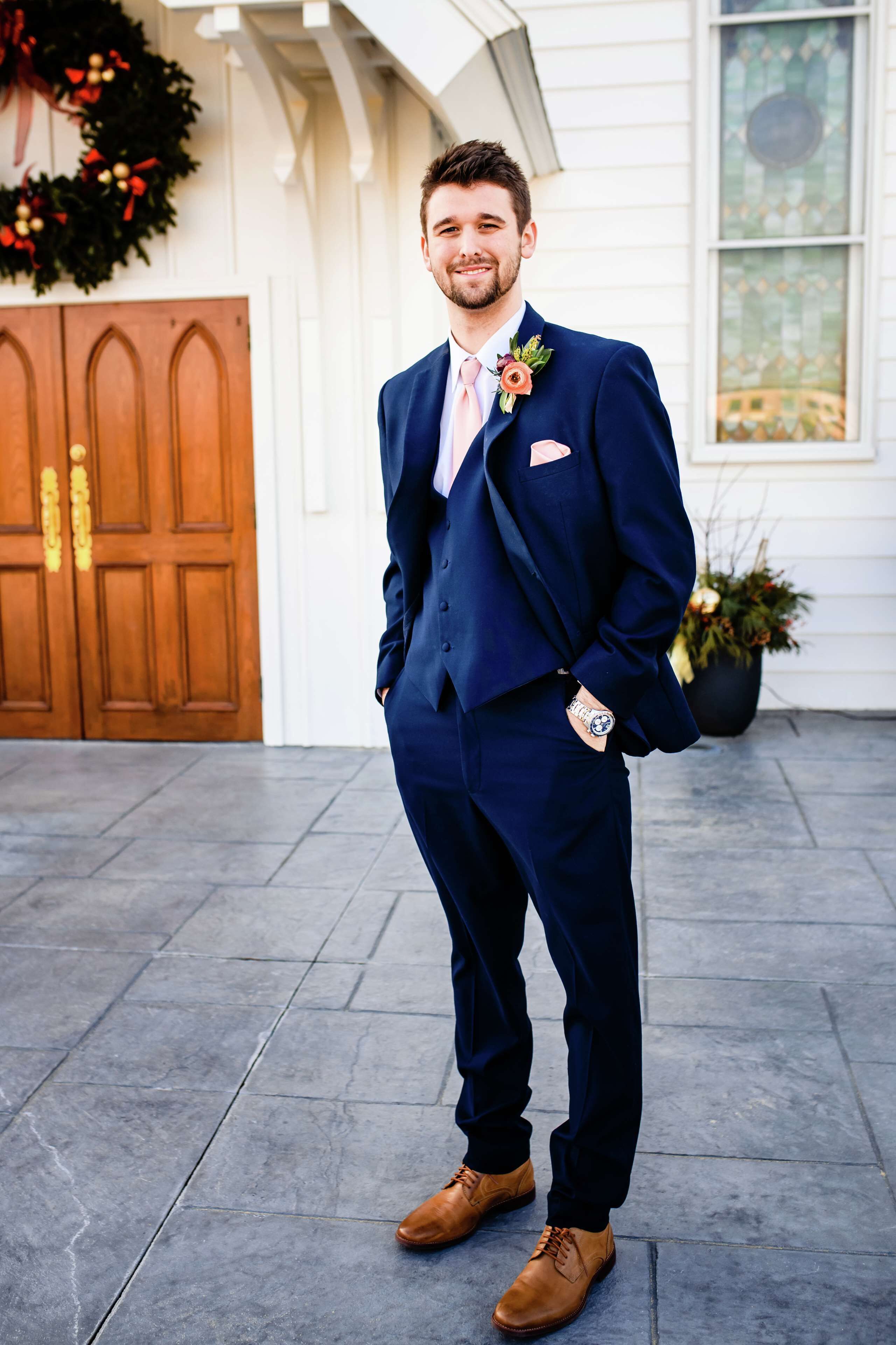 A man in a dark blue suit with a boutonniere, standing on a tiled floor with a wooden door and a wreath in the background.