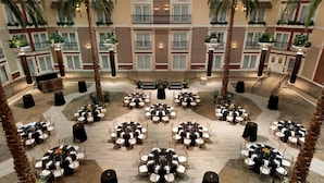 A hotel atrium with multiple round tables and chairs, palm trees, and balconies.