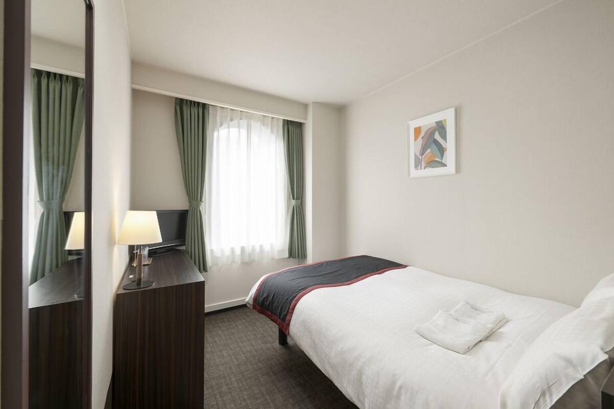 A hotel room with a bed, a desk with a lamp, a window with curtains, and a painting on the wall.