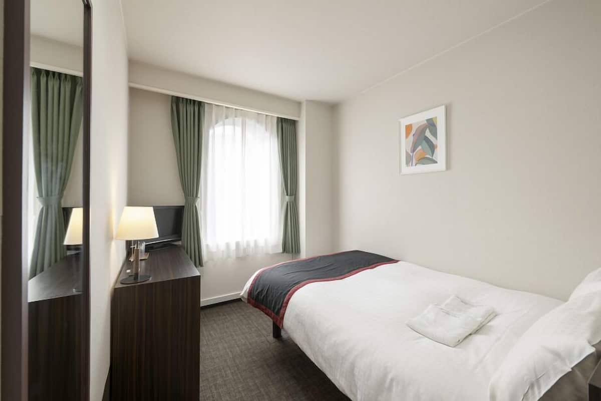 A hotel room with a bed, a desk with a lamp, a window with curtains, and a painting on the wall.