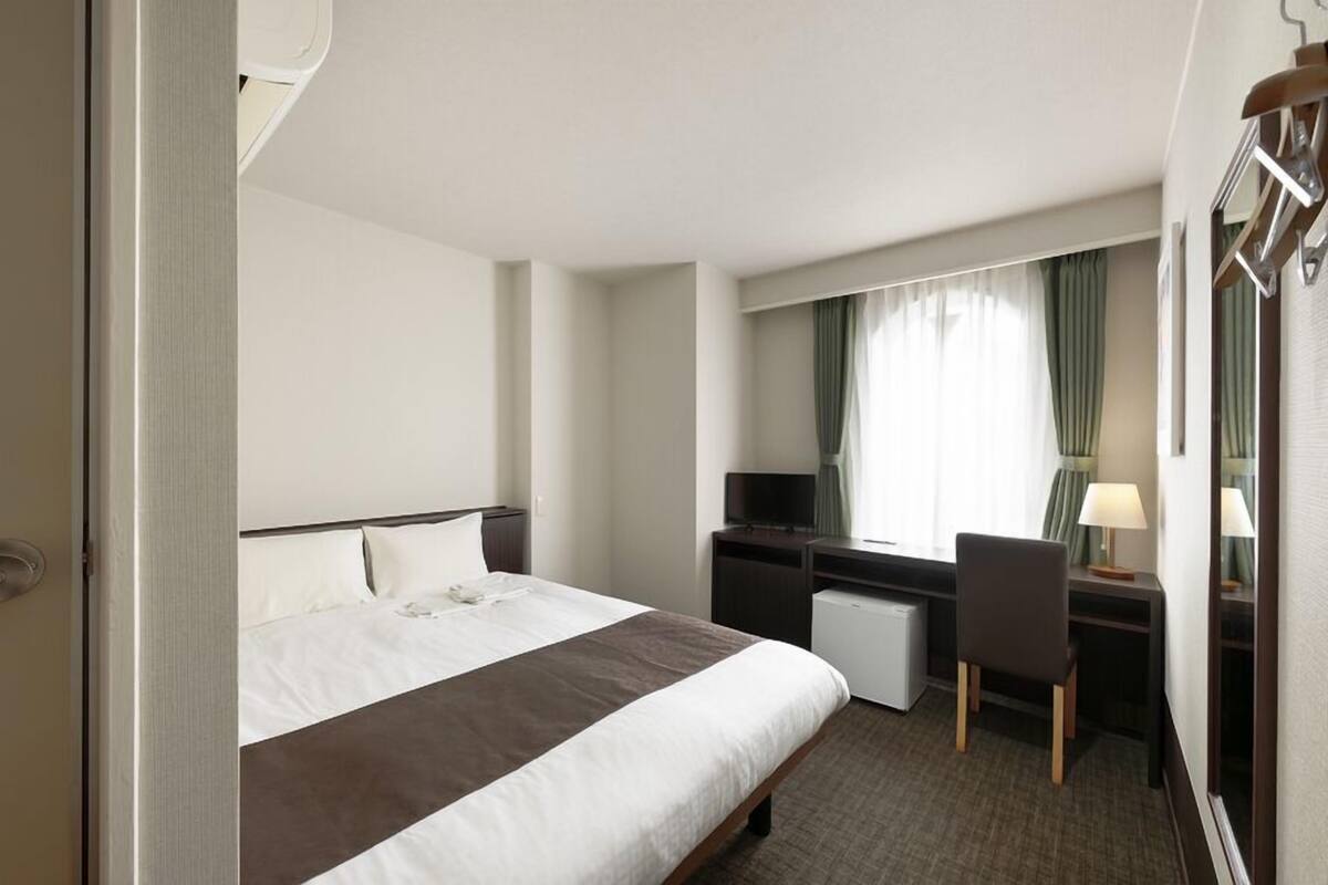 A hotel room with a bed, desk, chair, TV, and a window with curtains.