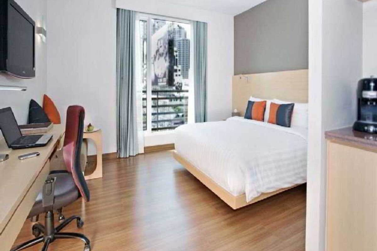 A hotel room with a bed, a desk with a laptop, a chair, a TV, and a window with a view of buildings.