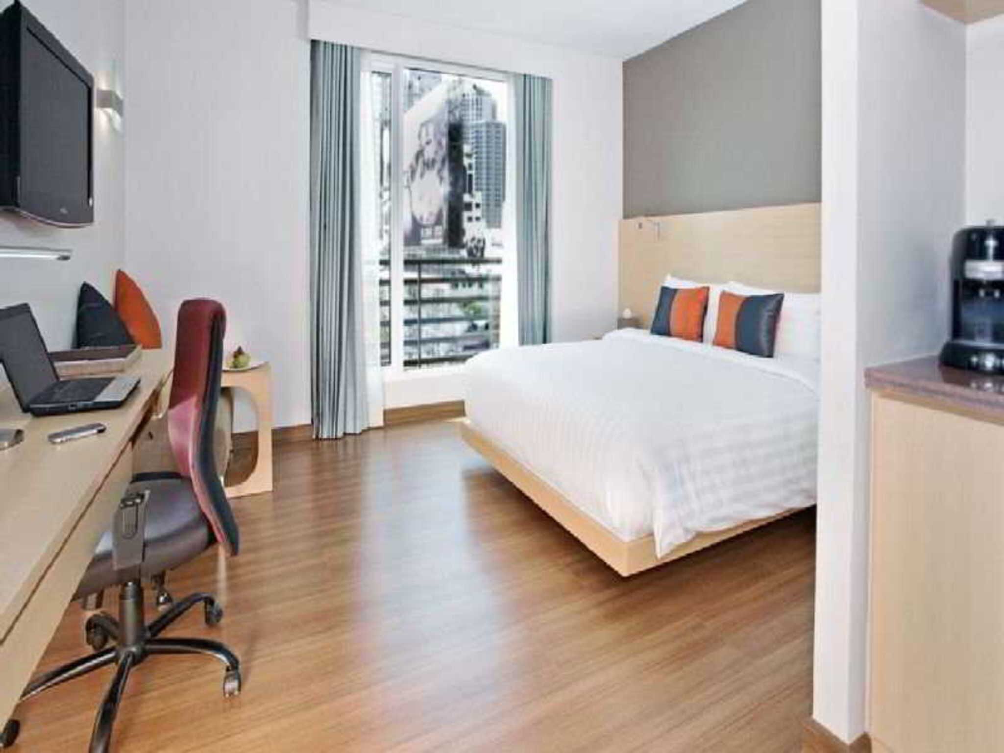 Une chambre d’hôtel avec un lit, un bureau avec un ordinateur portable, une chaise, une télévision et une fenêtre donnant sur des immeubles.