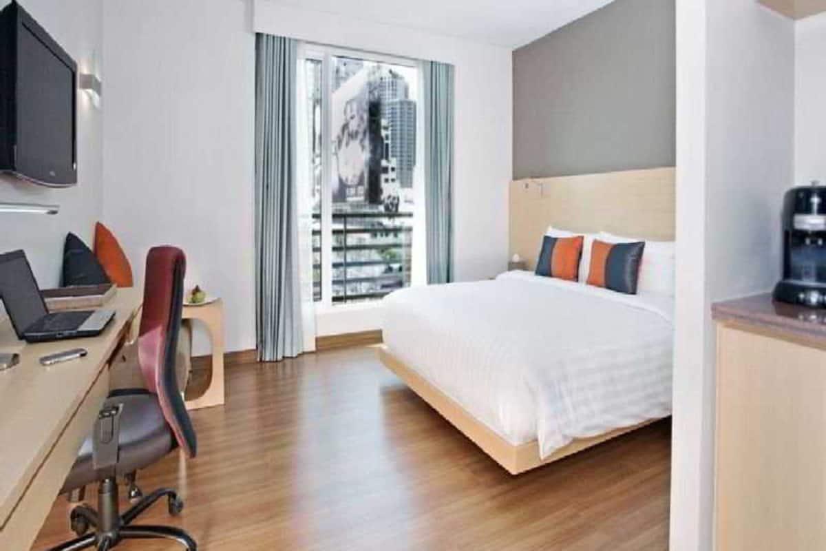 A hotel room with a bed, a desk with a laptop, a chair, a TV, and a window with a view of buildings.
