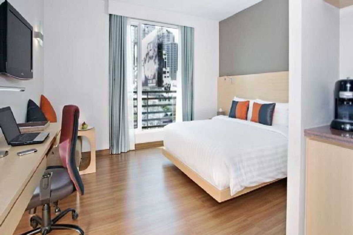 Une chambre d’hôtel avec un lit, un bureau avec un ordinateur portable, une chaise, une télévision et une fenêtre donnant sur des immeubles.