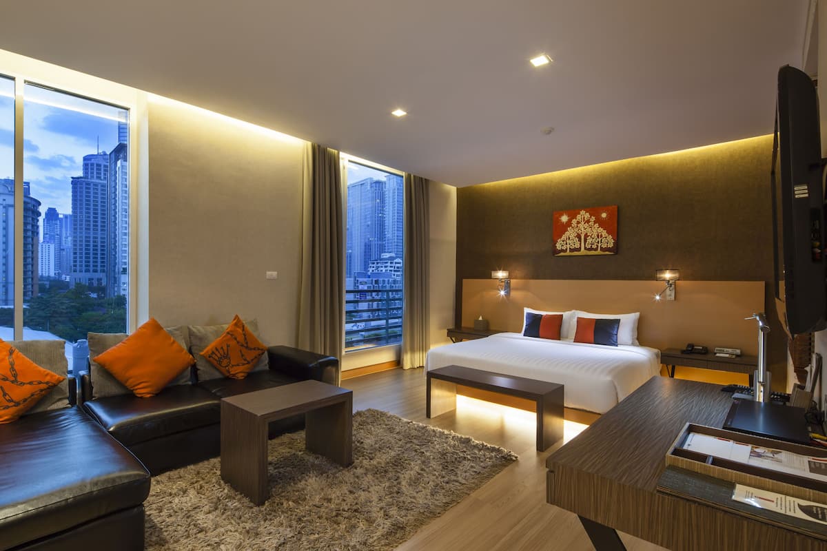 A modern hotel room with a large bed, a sofa, a desk, and a view of the cityscape through large windows.