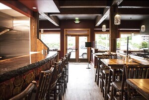 Un interior de restaurante con mesas y sillas de madera, una zona de bar y grandes ventanales.