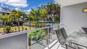 Un balcon avec une table et des chaises en verre, donnant sur une piscine et une végétation luxuriante.