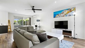 Un salon moderne avec un canapé d’angle, un téléviseur à écran plat et un tableau représentant une ville côtière.