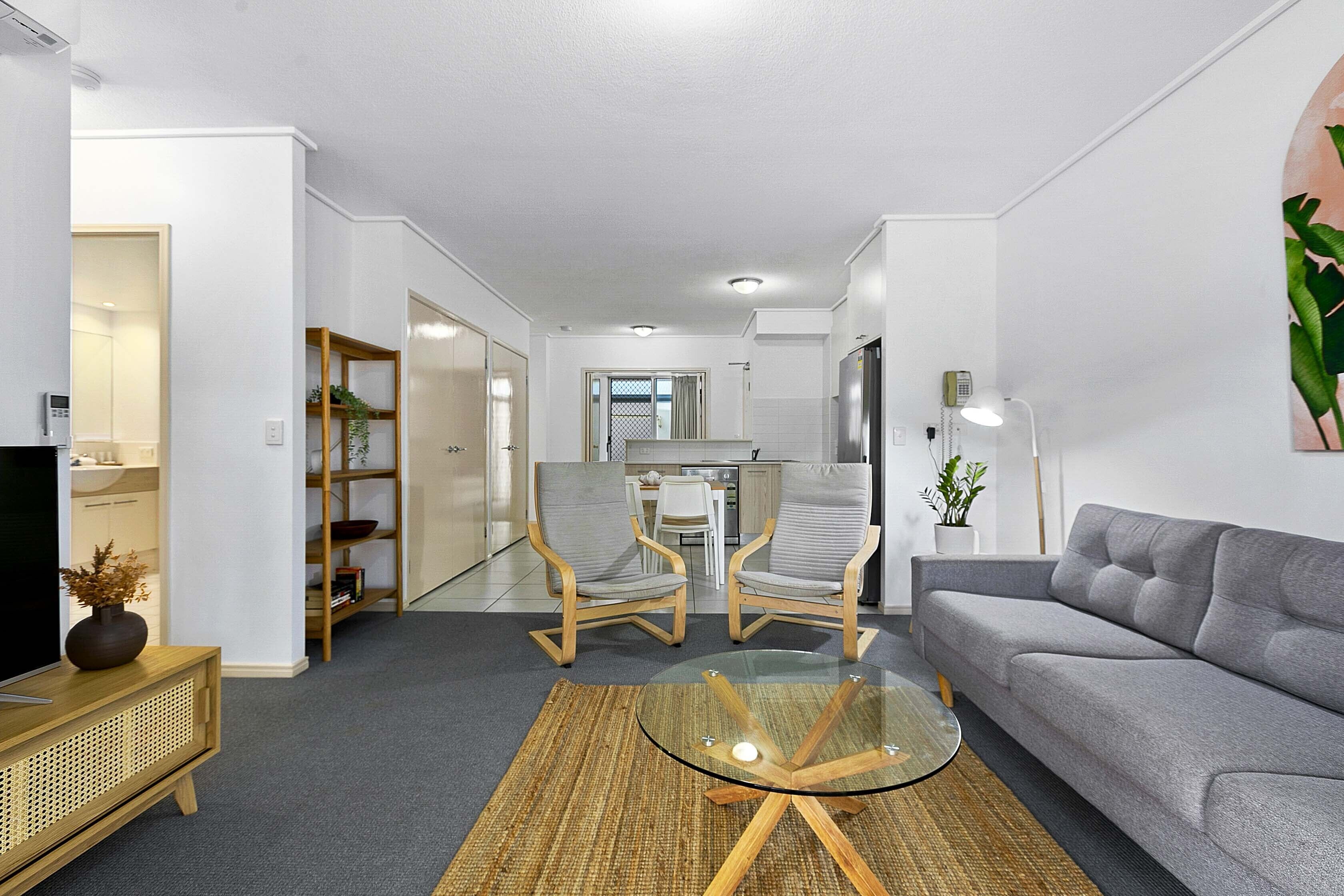A modern living room with a grey sofa, two wooden chairs, a glass coffee table, and a TV on a wooden cabinet.