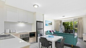 Une cuisine moderne avec des meubles blancs, un coin repas avec une table et des chaises, et un canapé vert dans un salon donnant sur un espace verdoyant.