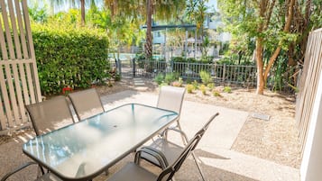 Une terrasse extérieure avec une table en verre et des chaises en métal, entourée d’un jardin où poussent différentes plantes, et une piscine en arrière-plan.