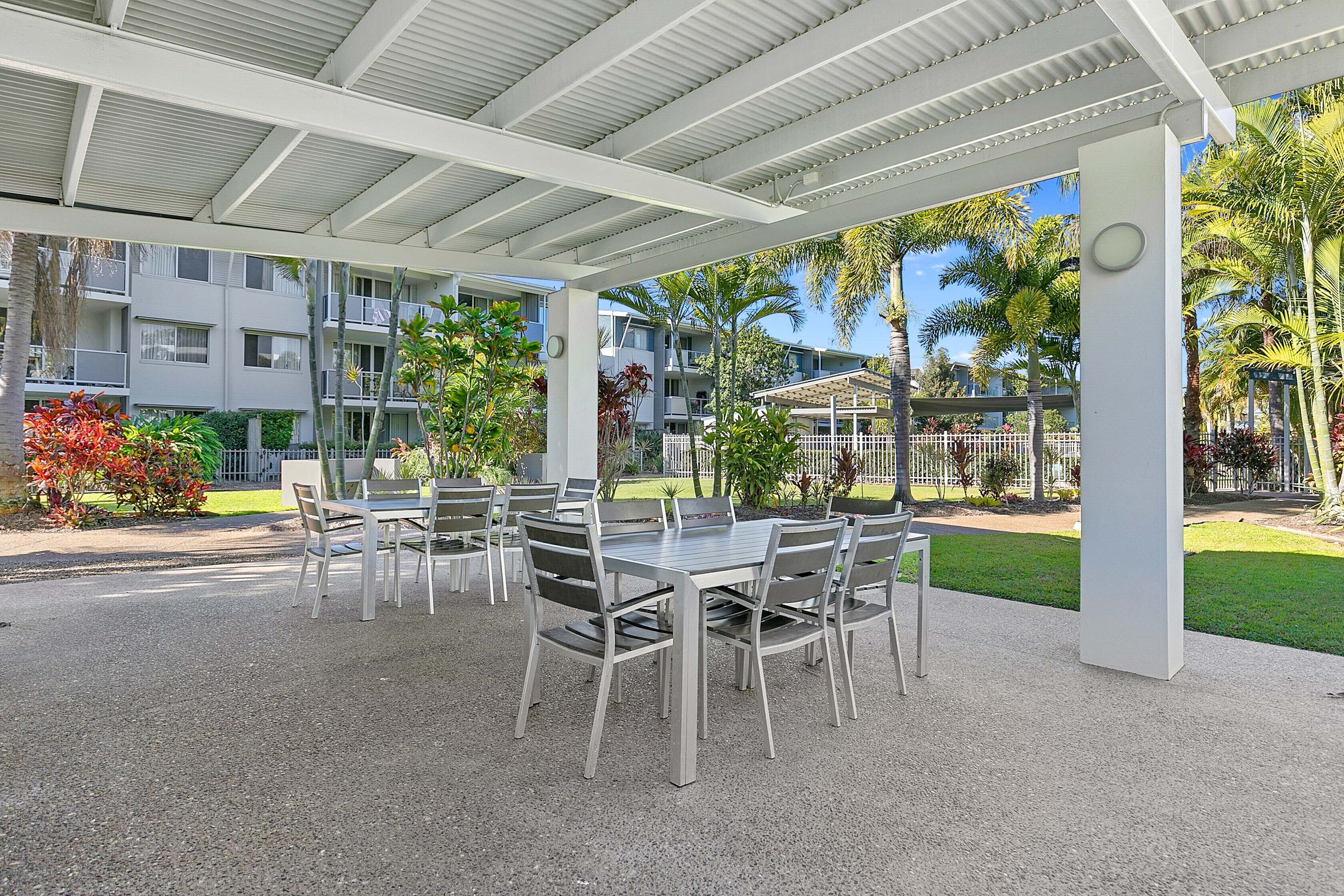 An outdoor patio with a covered seating area, surrounded by a well-maintained garden and residential buildings in the background.