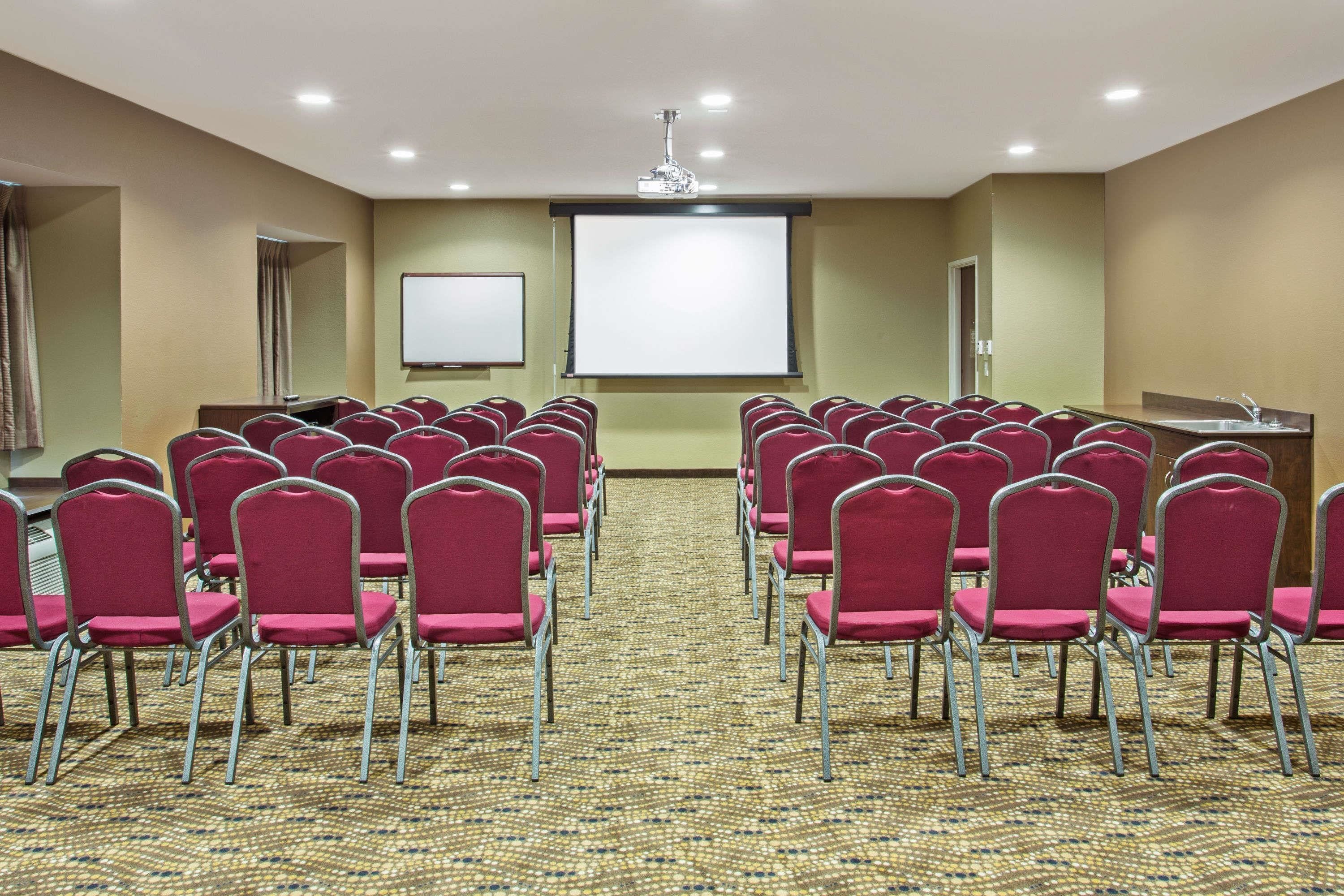 Un salón de conferencias con hileras de sillas rojas, una pantalla de proyector y suelo alfombrado.