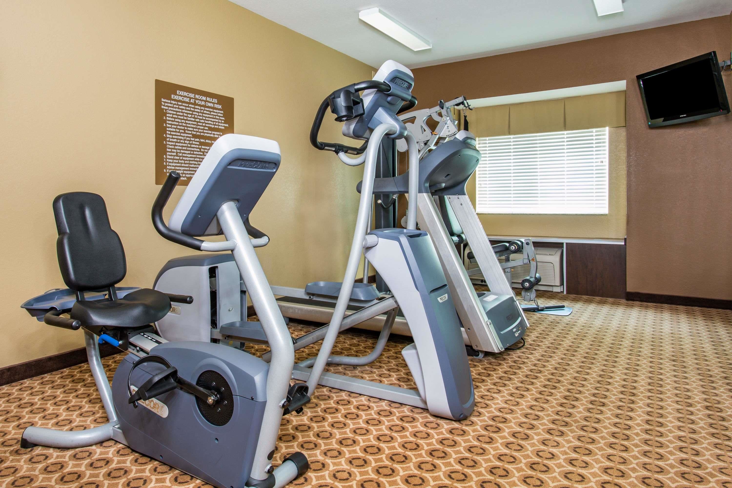 A fitness room with cardio machines, a wall-mounted TV, and a window with blinds.