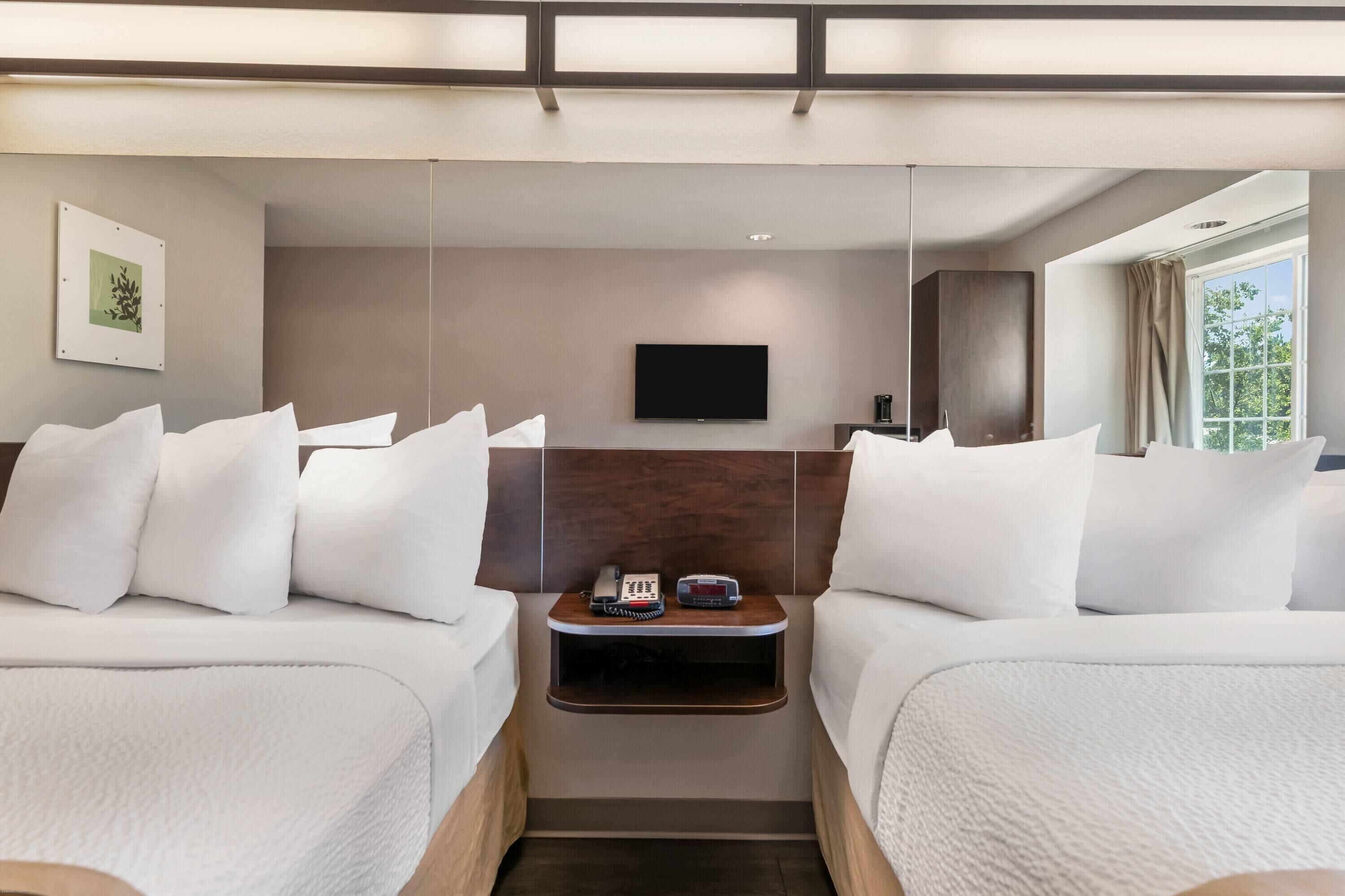A hotel room with two beds, a TV mounted on the wall, a small table with a phone and a coffee maker, and a window with curtains.