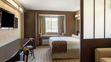 A hotel room with a bed, a desk with two chairs, a window with curtains, and a television mounted on the wall.