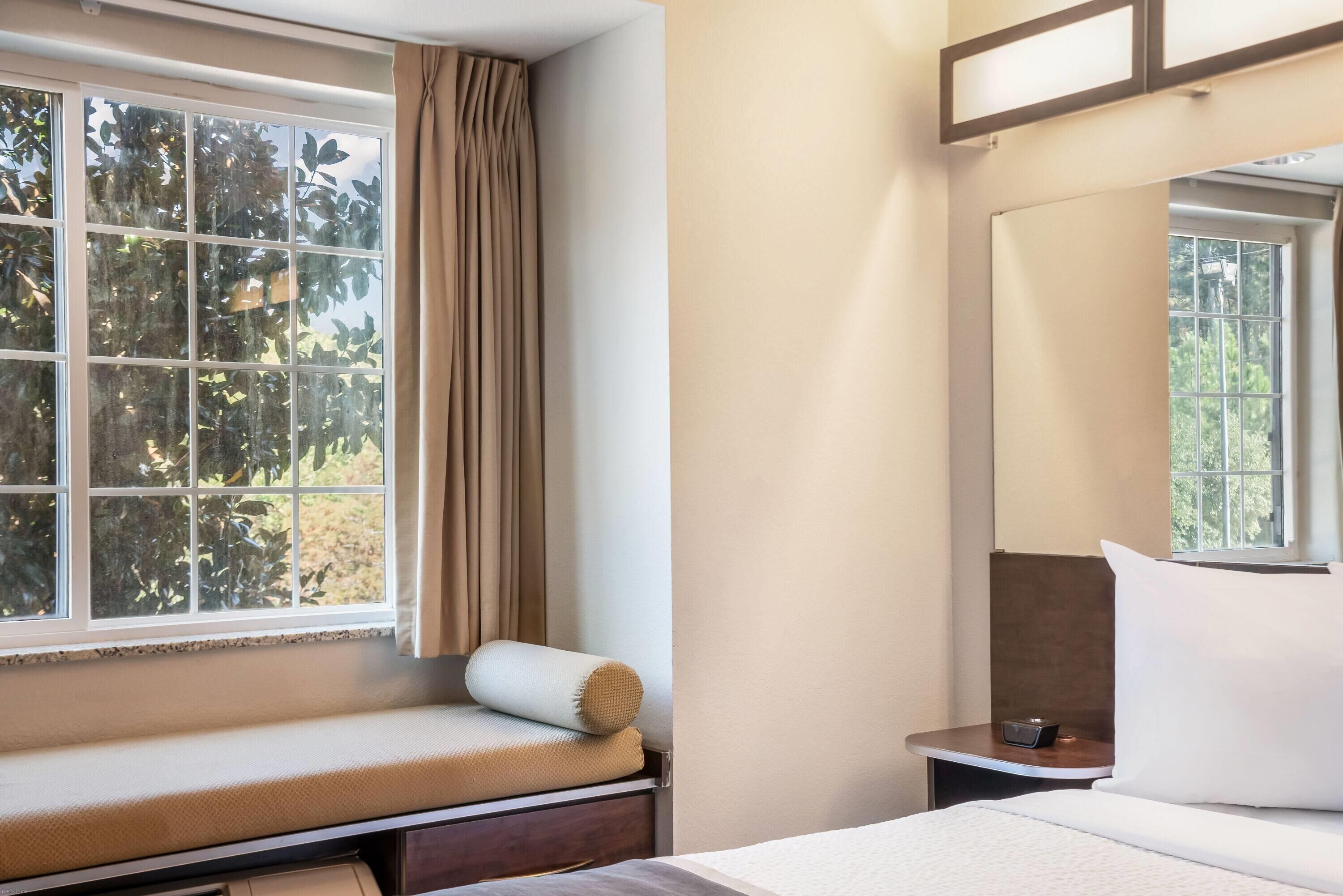 Une chambre d’hôtel avec un lit, une table de chevet, une fenêtre avec des rideaux et une vue sur les arbres à l’extérieur.