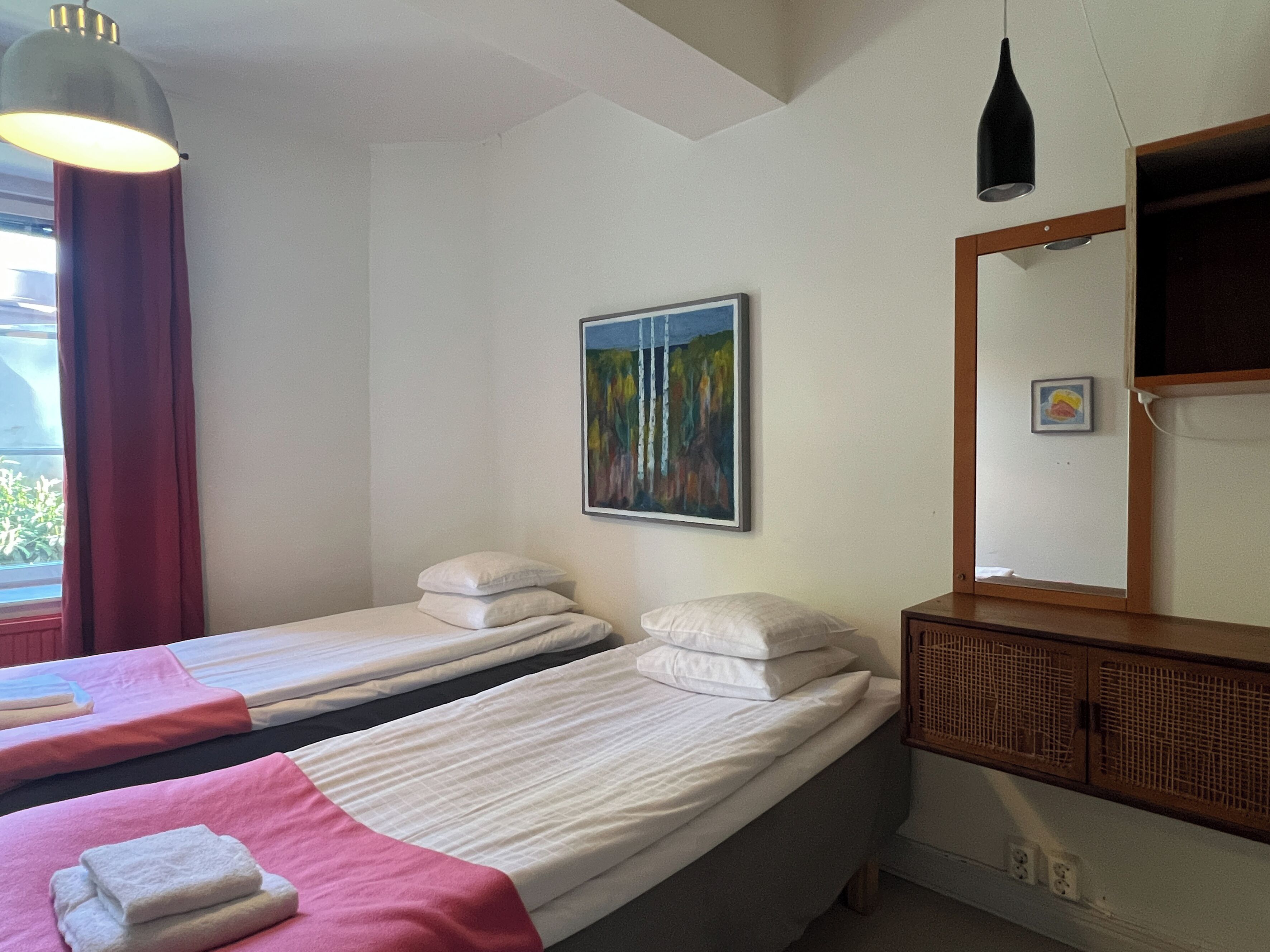 A hotel room with two single beds, a wooden dresser, a painting on the wall, and a window with curtains.