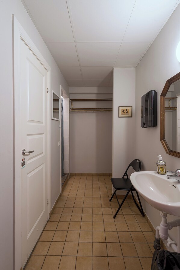 A narrow hallway with a white door, a tiled floor, a black chair, a white sink, and a mirror. - Reimersholme Hotel (Stockholm)
