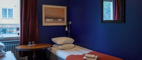 A small, neatly arranged room with a single bed, a small table, and a window with curtains.