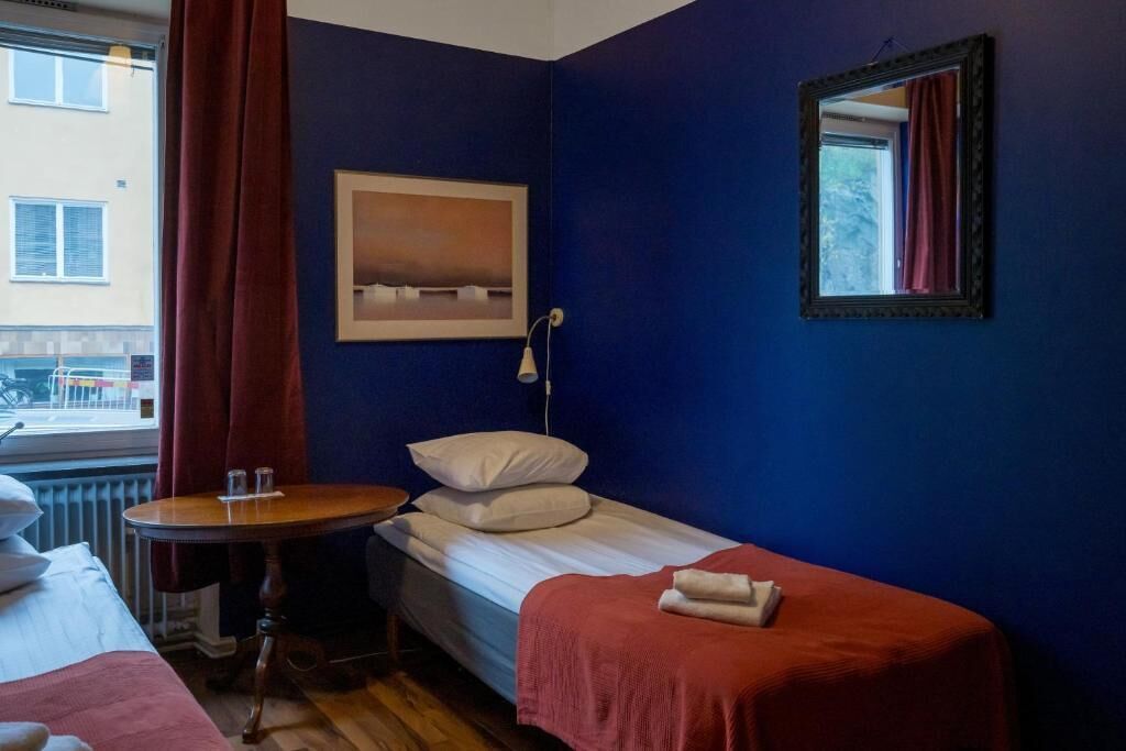 A small room with a single bed, a small table, a chair, a window with a view of buildings, and a framed picture on the wall.