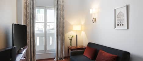 A modern hotel room with a flat-screen TV, a dark-colored sofa with red cushions, a side table with a lamp, and a framed picture on the wall.