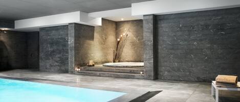 Un área de piscina interior moderna con una piscina azul cristalina, paredes de piedra oscura y un banco con una toalla.