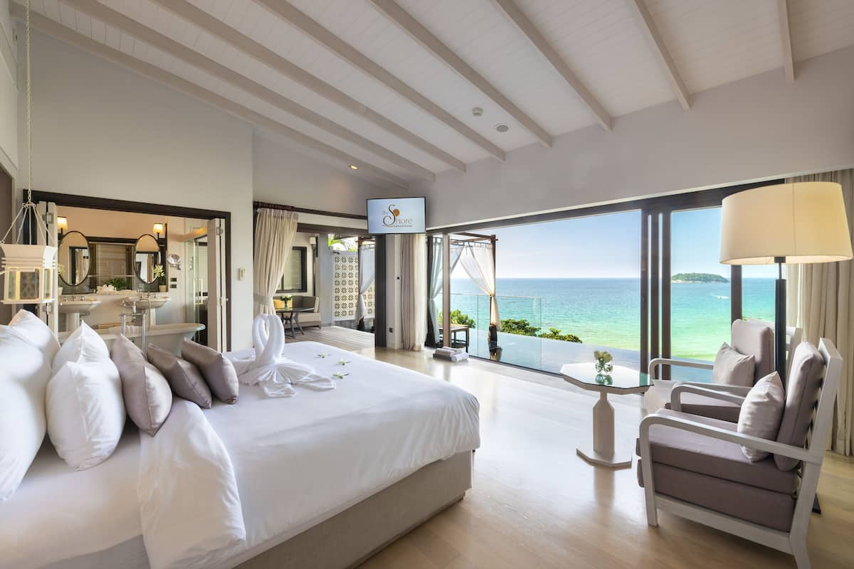 A spacious bedroom with a large bed, a sitting area with a view of the sea, and a bathroom with a vanity and mirror.