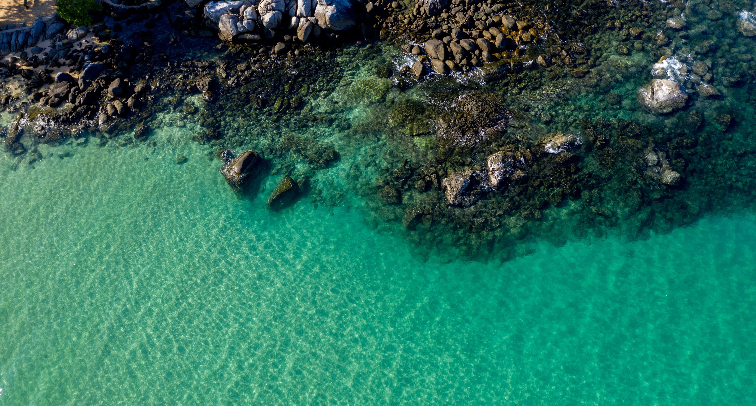 An aerial view of a coastal area with clear turquoise water, rocky shorelines, and large boulders.