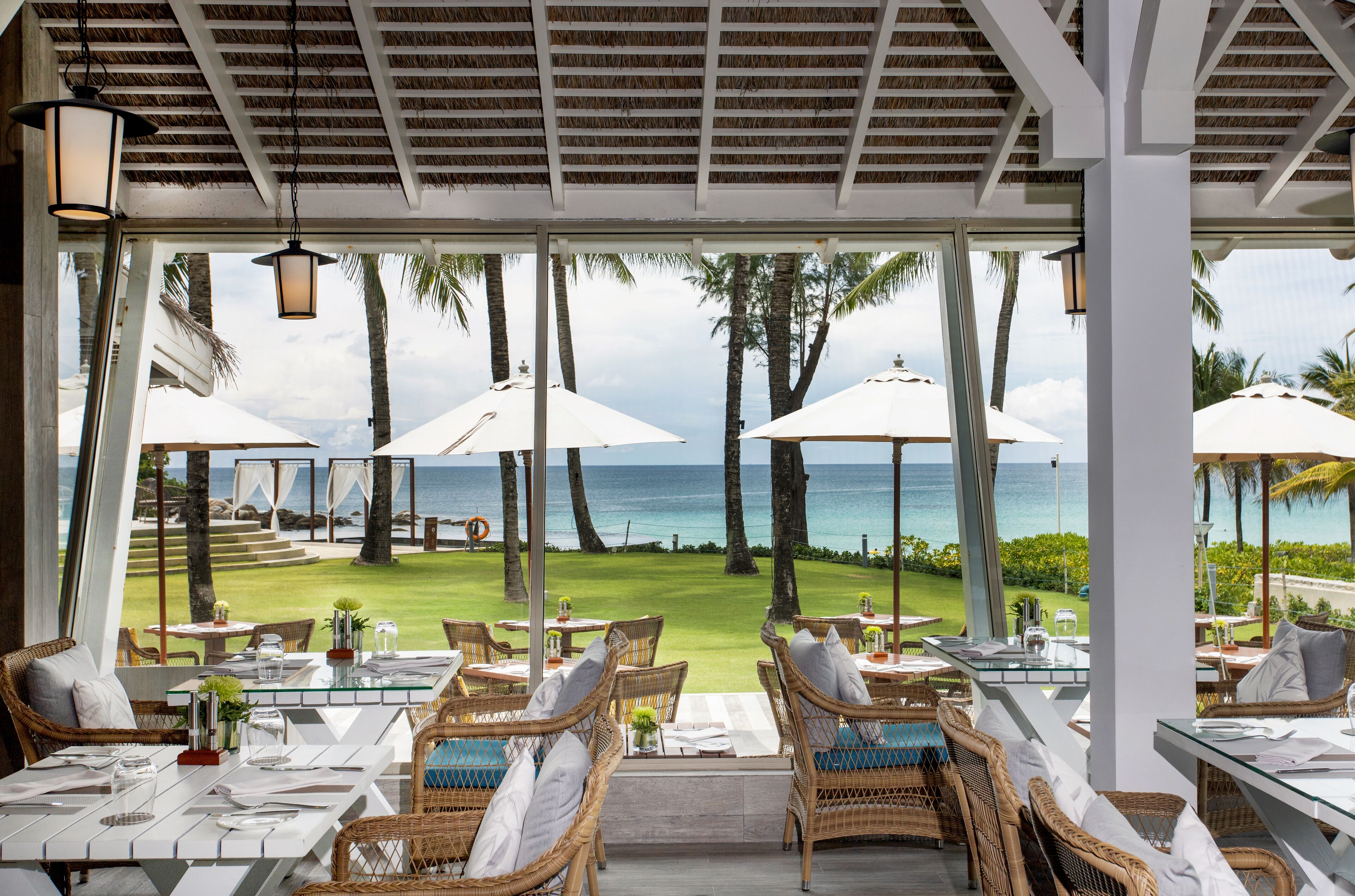 Un espace extérieur aménagé pour les repas, avec des meubles en osier, des tables blanches et des parasols, offrant une vue sur une plage bordée de palmiers et sur l’océan.