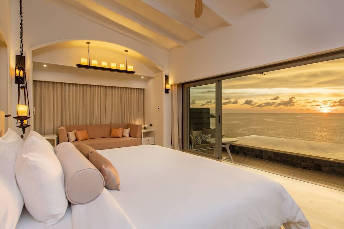 Ein geräumiges Schlafzimmer mit einem großen Bett, einer Couch und Blick auf den Ozean bei Sonnenuntergang.