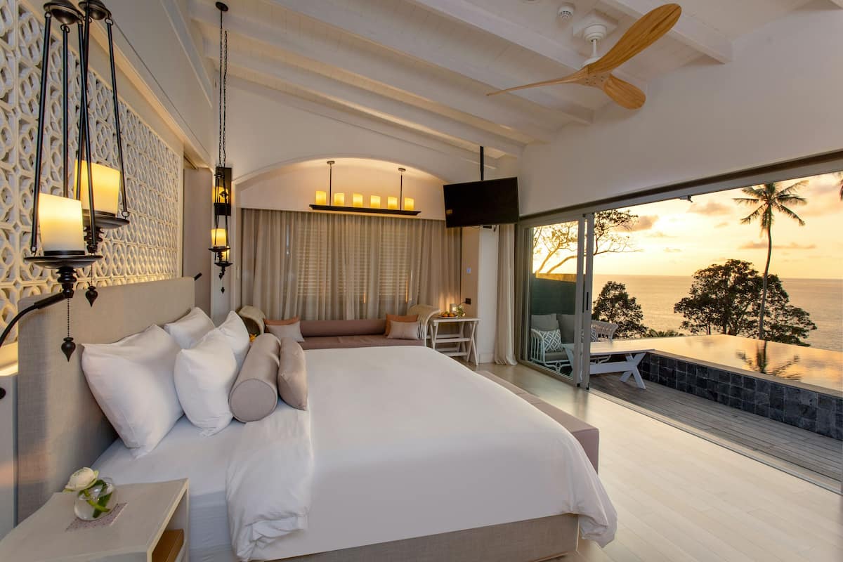 Ein geräumiges Schlafzimmer mit einem großen Bett, Deckenventilator und Blick auf das Meer bei Sonnenuntergang.