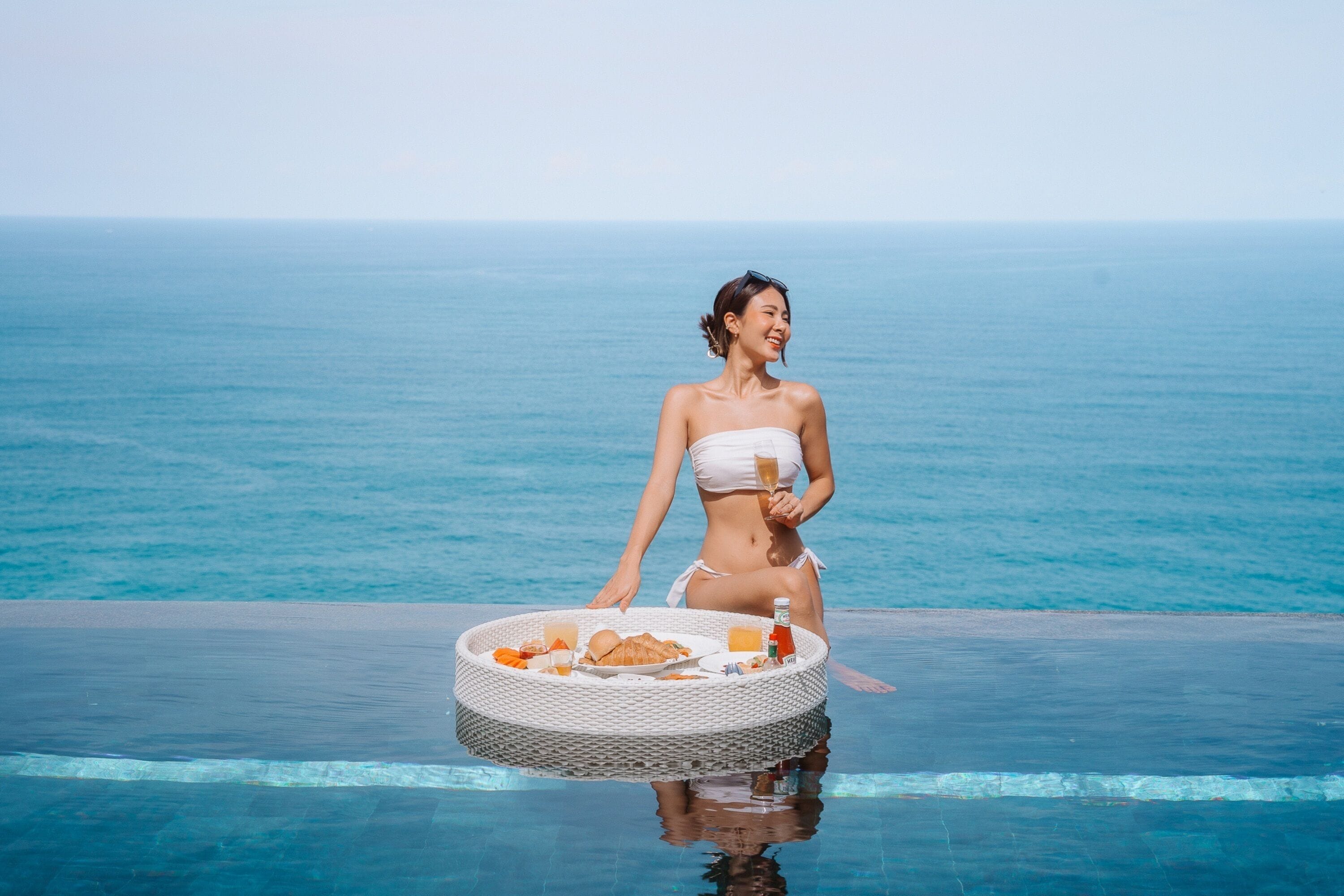 A person sitting by a pool with a tray of food and drinks, overlooking the ocean.