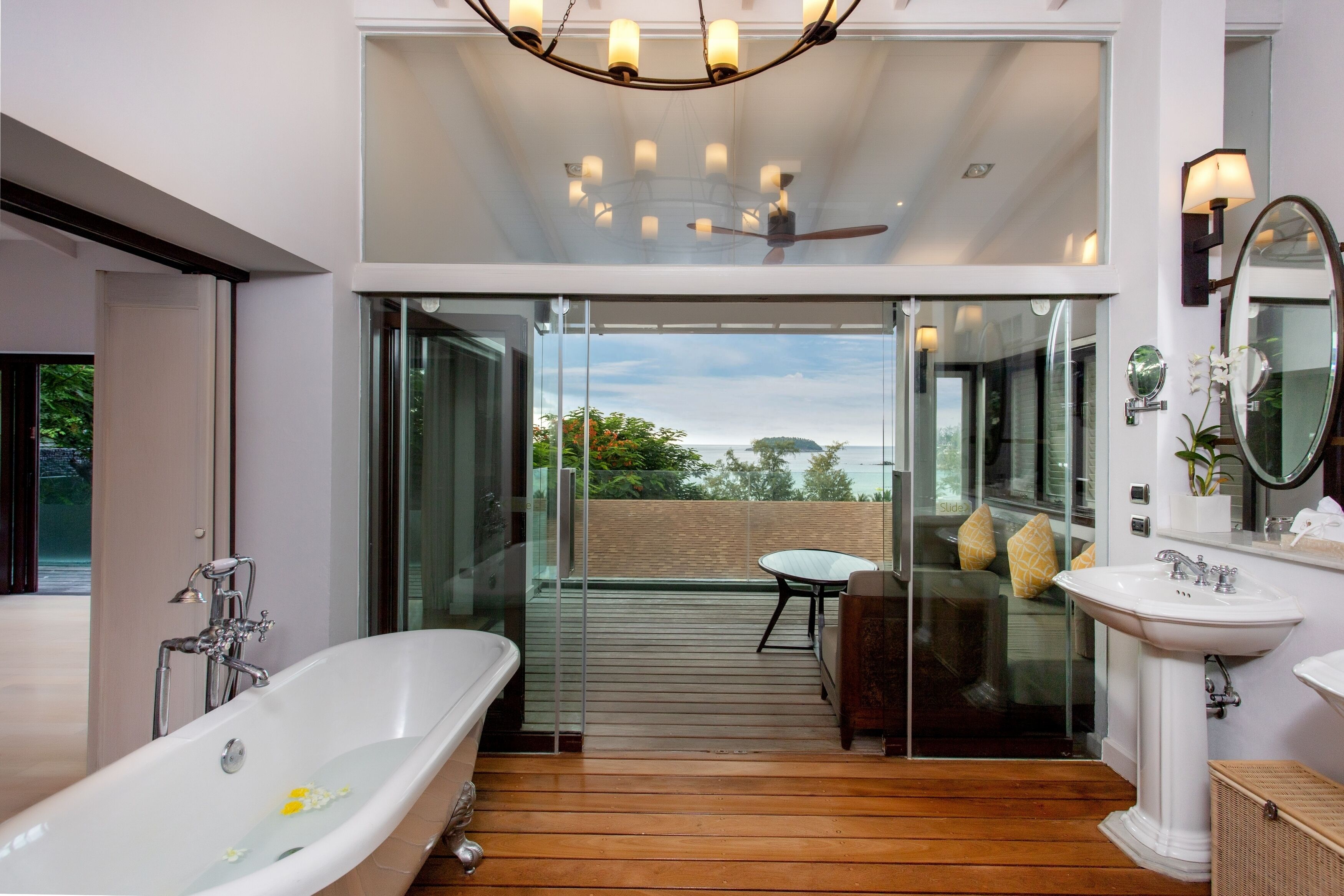 Un baño moderno con una bañera independiente, zona de ducha con mampara de vidrio y terraza de madera que conduce a un balcón con vista al mar.
