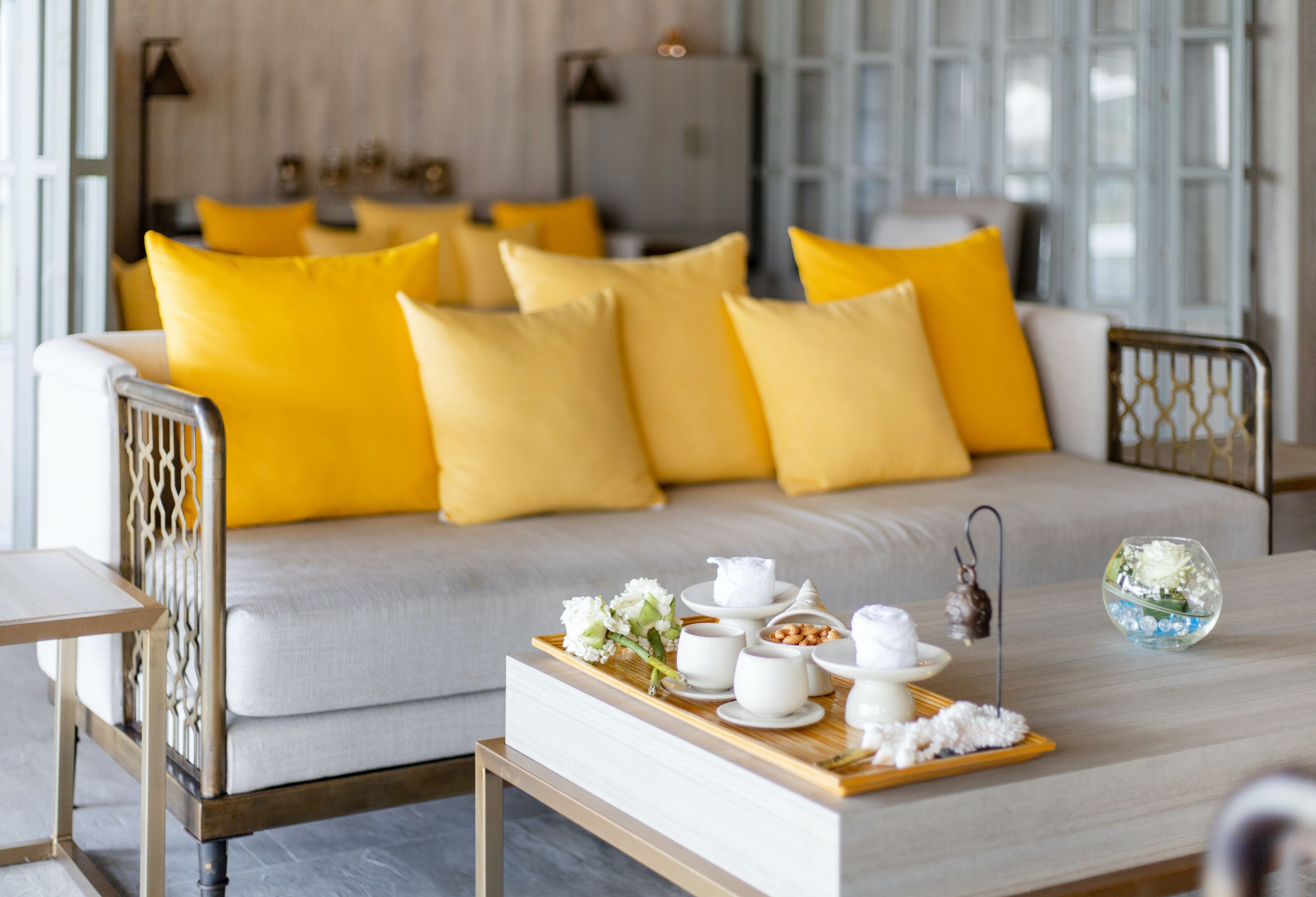A modern living room with a sofa adorned with yellow pillows, a coffee table with a tray of white teacups and a floral arrangement.