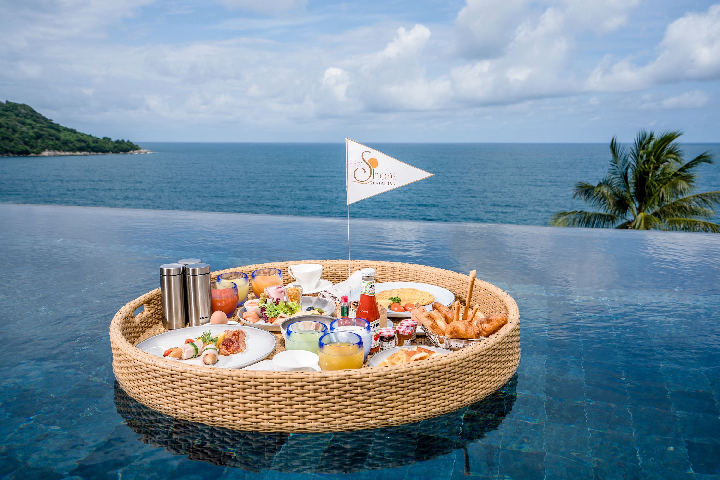 A floating breakfast setup with a flag, surrounded by a body of water with a view of the sea and a palm tree.