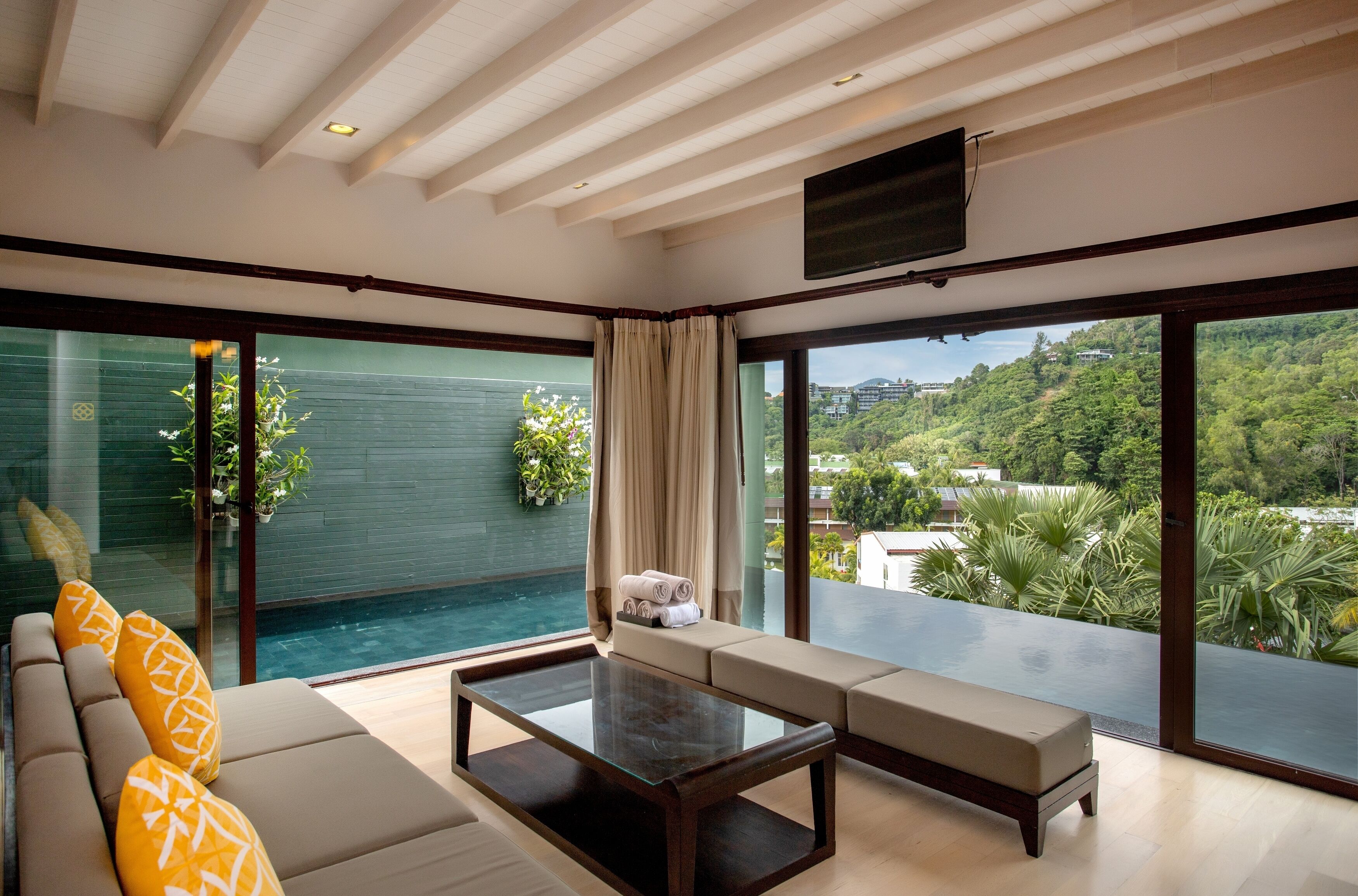 A modern living room with a glass wall, a sofa, a coffee table, and a view of a lush green landscape.